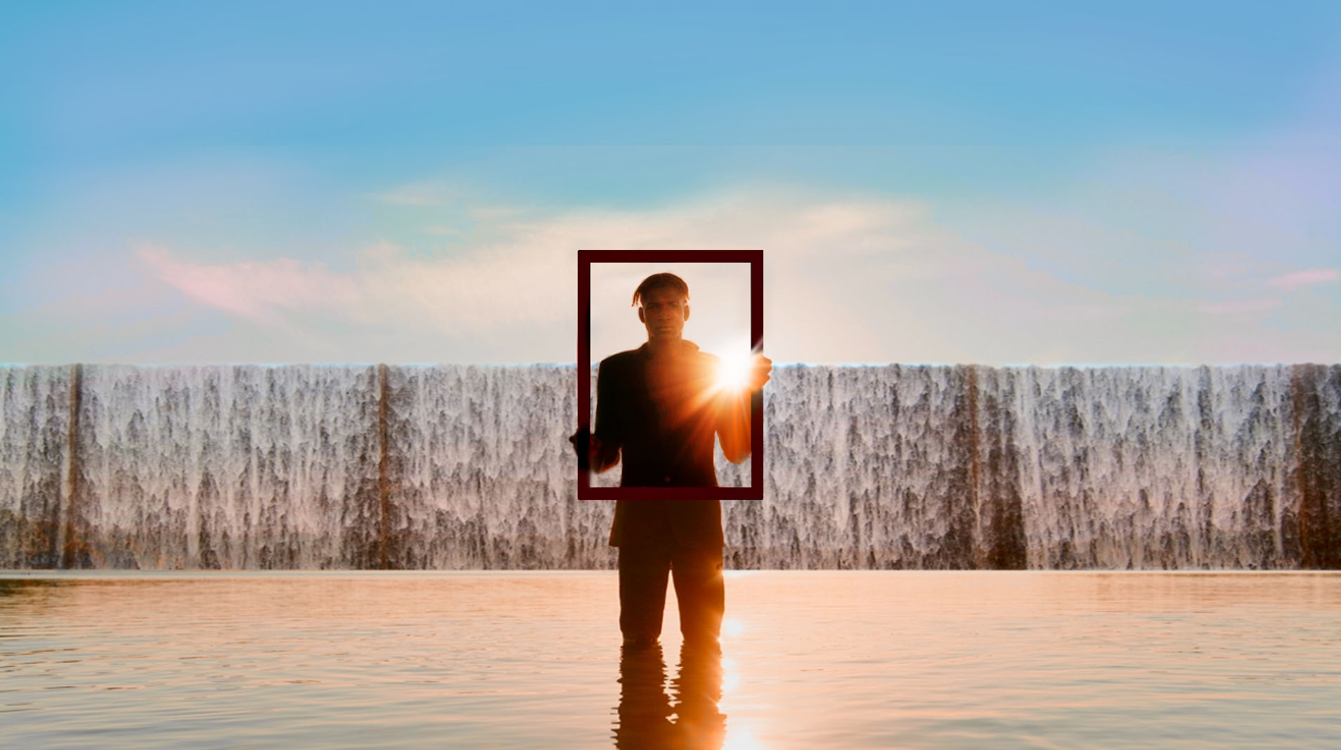 A person stands in shallow water holding an empty rectangular frame in front of their body, with a waterfall and bright sunlight in the background, creating a striking scene that feels like a landing between adventure and home.